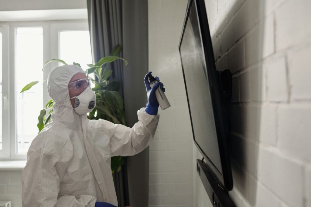 A person wearing full PPE disinfects a TV screen indoors, emphasizing health safety.