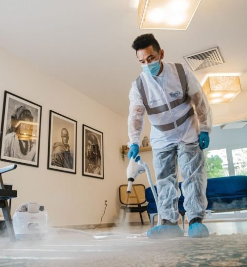 A professional in protective gear steam cleans carpet indoors, maintaining hygiene.