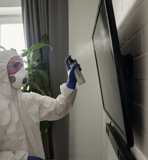 A person wearing full PPE disinfects a TV screen indoors, emphasizing health safety.