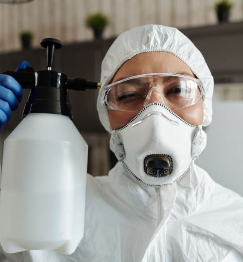Female wearing protective equipment and spraying disinfectant indoors.