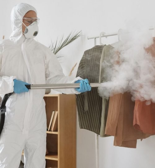 A person in PPE suit using a fumigation device to disinfect clothes indoors.